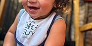 Micaden is registered to the contest to win money with this photo: baby, child, smiling, curly_hair, bib, blue_clothing, sitting, happy, closeup, face, skin, arm, foot, indoor, wooden_chair, blurred_background, portrait, cute, infant, natural_light