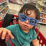 toddler, child, shopping_cart, sunglasses, striped_shirt, toy, indoor, store, aisle, curly_hair, reaching_hand, red_cart, shopping, baby, retail, casual_clothing, cute, person, playful, fun