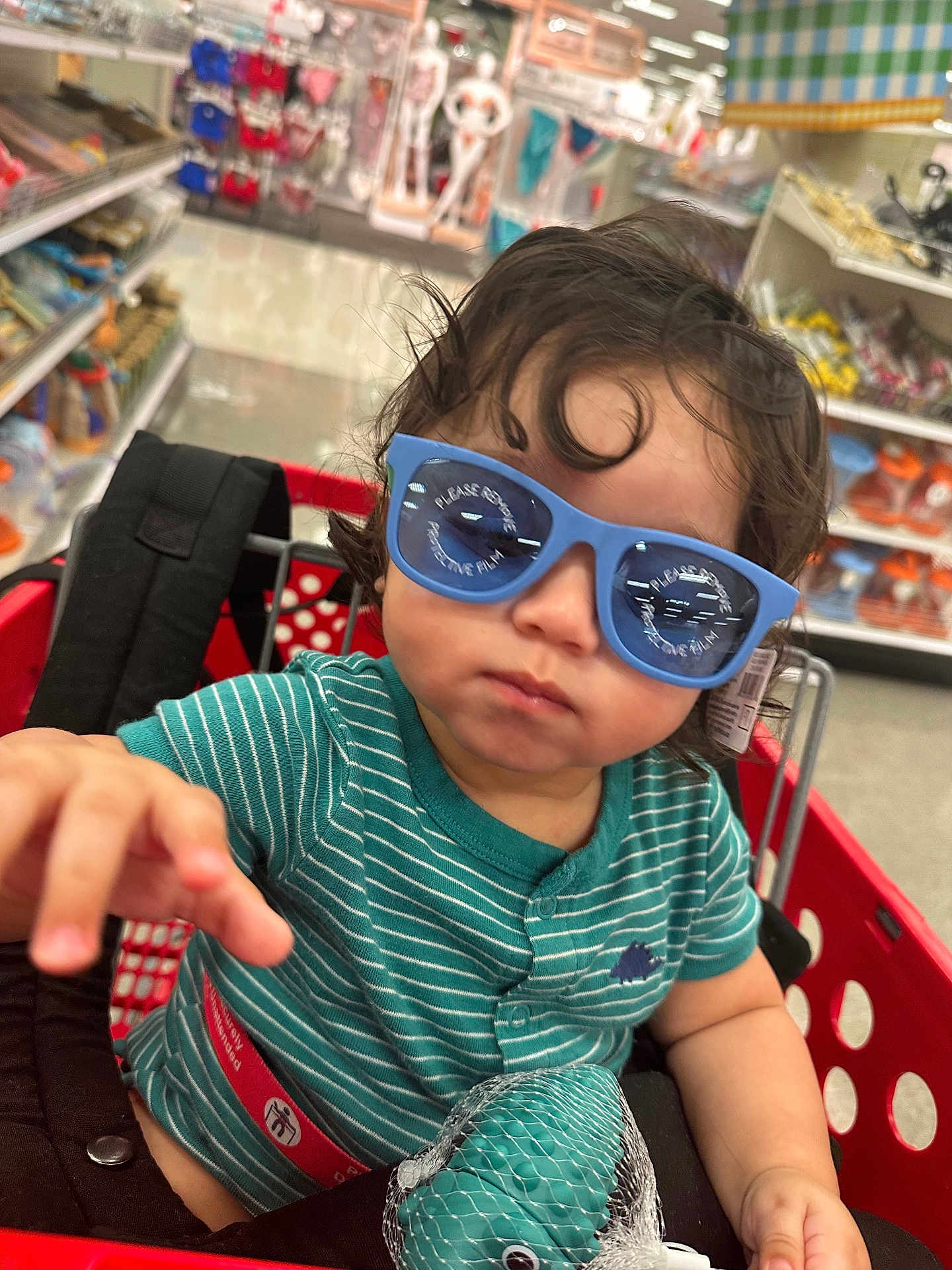 Micaden is registered to the contest to win money with this photo: toddler, child, shopping_cart, sunglasses, striped_shirt, toy, indoor, store, aisle, curly_hair, reaching_hand, red_cart, shopping, baby, retail, casual_clothing, cute, person, playful, fun
