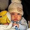 baby, infant, child, portrait, face, looking_at_camera, serious_expression, winter_hat, pom_pom, beanie, blanket, plush_toy, stuffed_animal, car_seat, cozy, bundled, clothing, indoor, close_up, photography