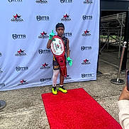 Ryan joined the competition — help win amazing prizes! child, red_carpet, championship_belt, sportswear, gloves, neon_cleats, cap, outdoor, concrete, banner, logo, pose, victory, young_athlete, celebration, portrait, event, person, background, photography