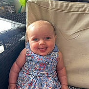 Charlotte is registered to the contest to win money with this photo: baby, child, smiling, tongue_out, blue_dress, floral_pattern, cushion, chair, indoor, happy, cute, person, infant, seated, plush, skin, face, head, bright_eyes, short_hair