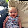baby, child, smiling, tongue_out, blue_dress, floral_pattern, cushion, chair, indoor, happy, cute, person, infant, seated, plush, skin, face, head, bright_eyes, short_hair