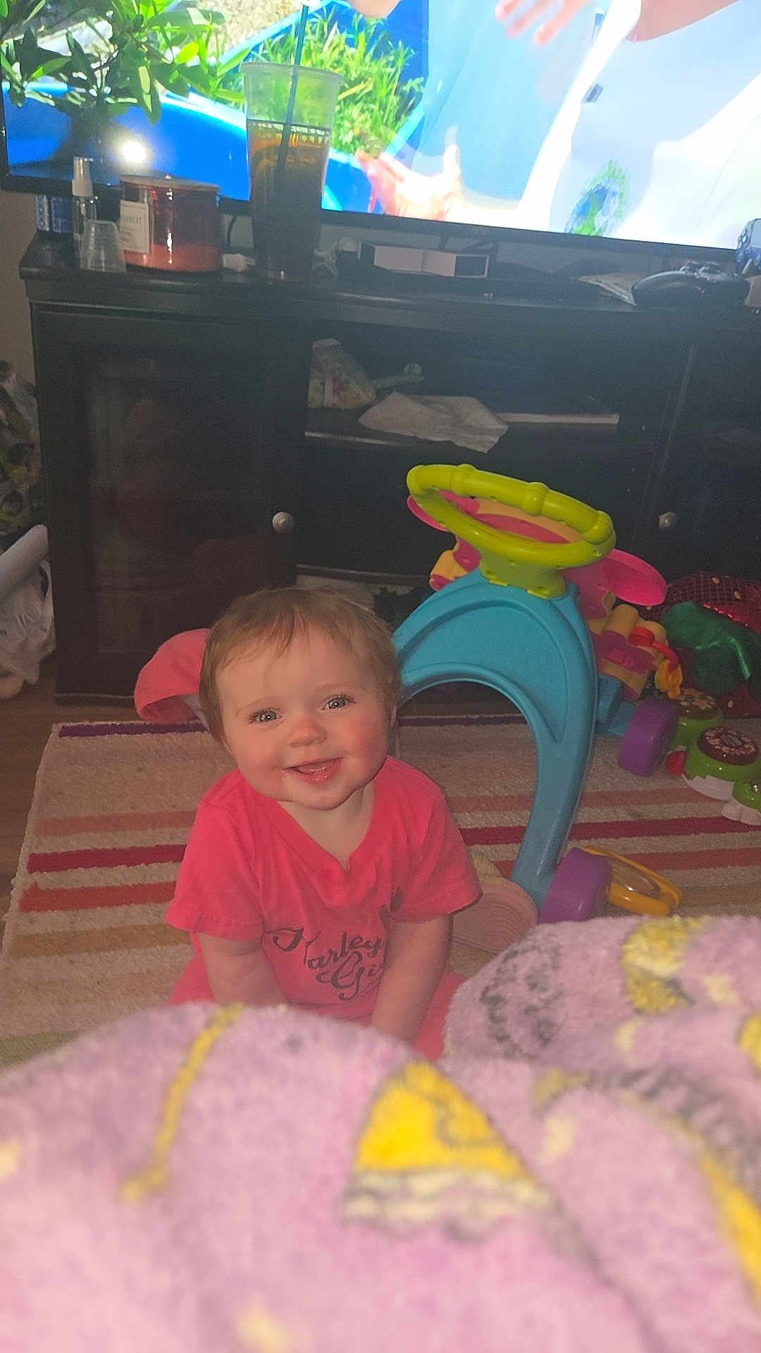 Charlotte is registered to the contest to win money with this photo: child, toddler, smiling, indoor, living_room, rug, blanket, television, cabinet, toy, ride_on_toy, floor, shirt, happy, furniture, cup, glass, candle, play, carpet