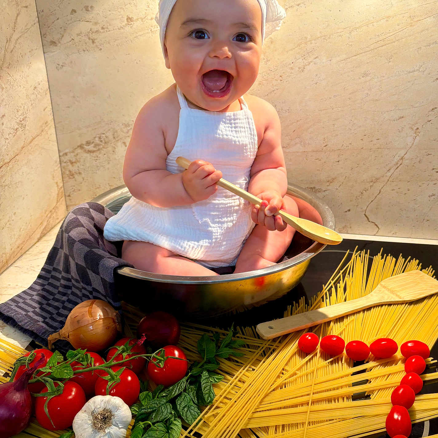 Leya a rejoint le concours — aidez-le/la à gagner de superbes lots ! baby, basil, child, cooking, food, fresh_ingredients, garlic, happy, headwrap, indoors, kitchen, metal_bowl, onion, playful, sitting, smiling, spaghetti, tomatoes, white_clothing, wooden_spoon