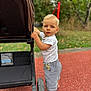 toddler, child, barefoot, stroller, outdoor, playground, grass, greenery, shorts, white_shirt, blue_eyes, curious, summer, daylight, person, walking, standing, handle, wheel, park