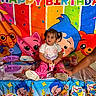 animal_theme, balloons, birthday, birthday_party, cake_stand, cartoon_characters, celebration, child, colorful, cupcake, decorations, indoors, party_table, pigtails, pink_pants, portrait, sitting, snack, toddler, white_shirt