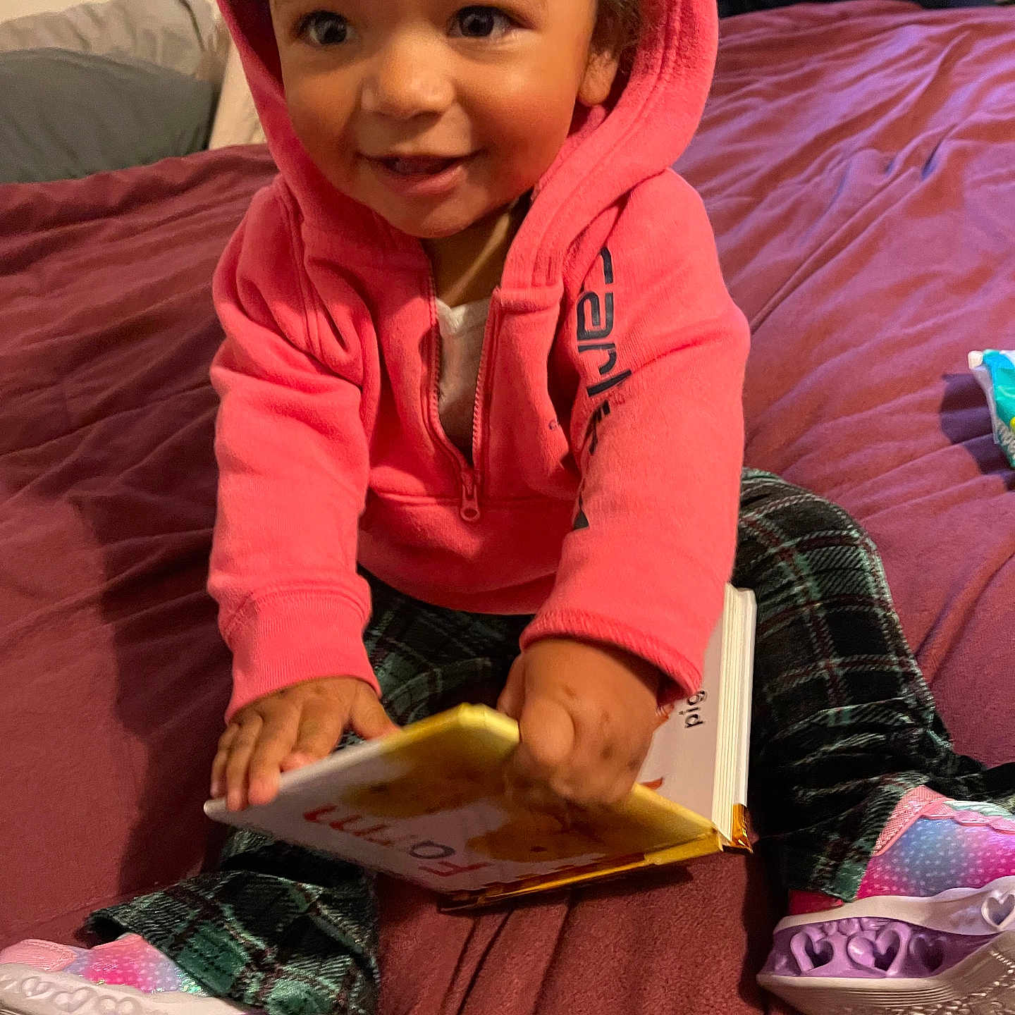 Calliah is registered to the contest to win money with this photo: bed, book, casual_clothing, child, colorful, cozy, cute, happy, hoodie, indoor, person, plaid_pants, playful, portrait, reading, shoes, sitting, smiling, toddler, young_child