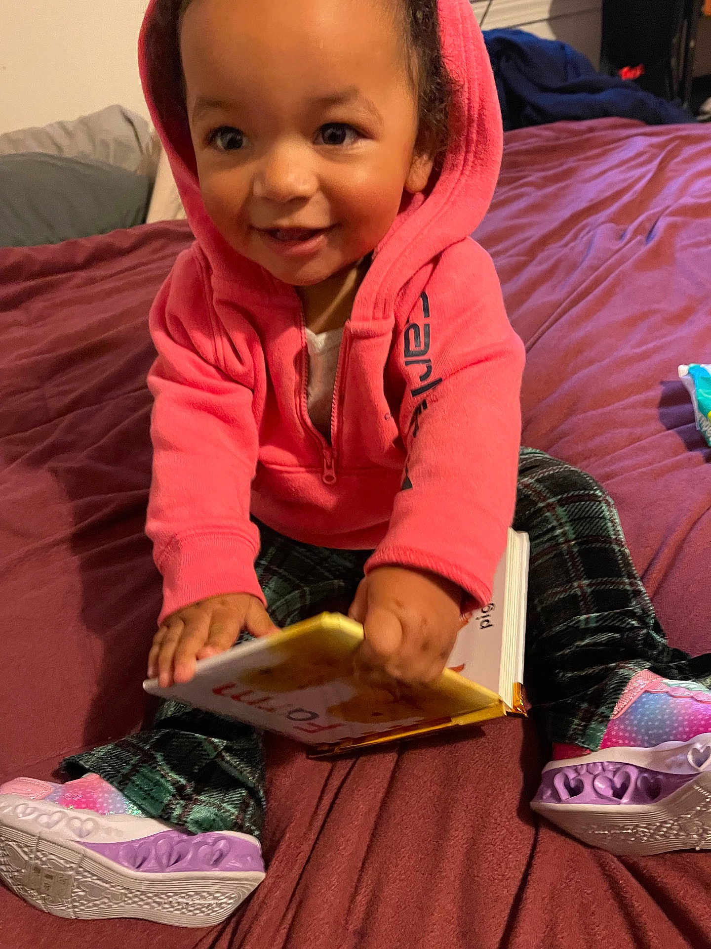 Calliah is registered to the contest to win money with this photo: toddler, child, smiling, hoodie, book, bed, indoor, happy, shoes, plaid_pants, person, young_child, sitting, reading, colorful, casual_clothing, cute, portrait, playful, cozy
