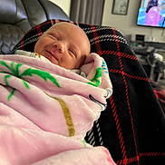 Noelle joined the competition — help win amazing prizes! baby, blanket, smiling, indoor, couch, living_room, television, plaid, face, person, child, cozy, comfort, home, soft, pink, green, black, red, wrapped