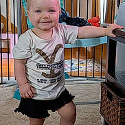 Taivyn is registered to the contest to win money with this photo: toddler, child, indoors, standing, pigtails, smiling, yellowstone_tshirt, black_shorts, barefoot, carpet, table, baby_gate, basket, wooden_door, toy, home, person, cute, balance, happy