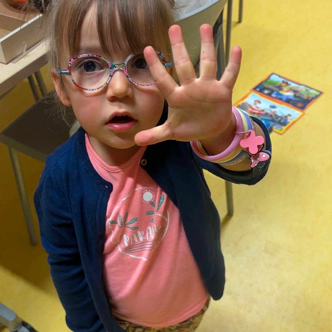 Léa a rejoint le concours — aidez-le/la à gagner de superbes lots ! bracelet, cardigan, chair, child, colorful, curious, expression, floor, floral_pants, glasses, hand, indoor, person, pink_shirt, playroom, puzzle, sandals, table, toy, young