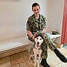 dog, husky, person, military_uniform, soldier, seated, indoor, bench, tiled_floor, boots, leash, military_gear, camouflage, portrait, animal, pet, tongue_out, fluffy, smiling, wall