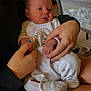 baby, newborn, infant, person, hands, clothing, onesie, giraffe, indoor, crib, furniture, nail_polish, holding, sitting, legs, footwear, television, blanket, home, casual