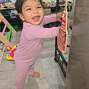 Aarin is registered to the contest to win money with this photo: toddler, child, pink_onesie, barefoot, smiling, indoor, wooden_floor, toys, furniture, bottle_opener, pirate, happy, standing, black_metal_shelf, carpet, curly_hair, home, playful, colorful, cute