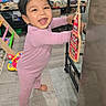 toddler, child, pink_onesie, barefoot, smiling, indoor, wooden_floor, toys, furniture, bottle_opener, pirate, happy, standing, black_metal_shelf, carpet, curly_hair, home, playful, colorful, cute