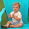 toddler, child, curly_hair, smiling, clapping, sitting, chair, adirondack_chair, blue_chair, outdoor, grass, casual_clothing, tshirt, shorts, sandals, summer, happy, playful, cute, portrait