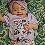 baby, infant, smile, purple_headwrap, bow, bracelet, patterned_bedspread, clothing, text_on_shirt, cute, lying_down, happy, child, portrait, pink_pants, hand, face, closeup, toddler, indoors
