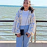 blue_pants, casual_clothing, crochet_jacket, crossbody_bag, female, hands, ocean, outdoor, person, phone, pier, portrait, railing, sea, smile, standing, sunlight, teenager, white_top, wind