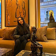 Rania a rejoint le concours — aidez-le/la à gagner de superbes lots ! young_woman, indoor, couch, sculpture, painting, window, building, gloves, jacket, long_hair, art, decor, brown_couch, pillows, marble_table, portrait, seated, relaxed, modern_interior, daylight