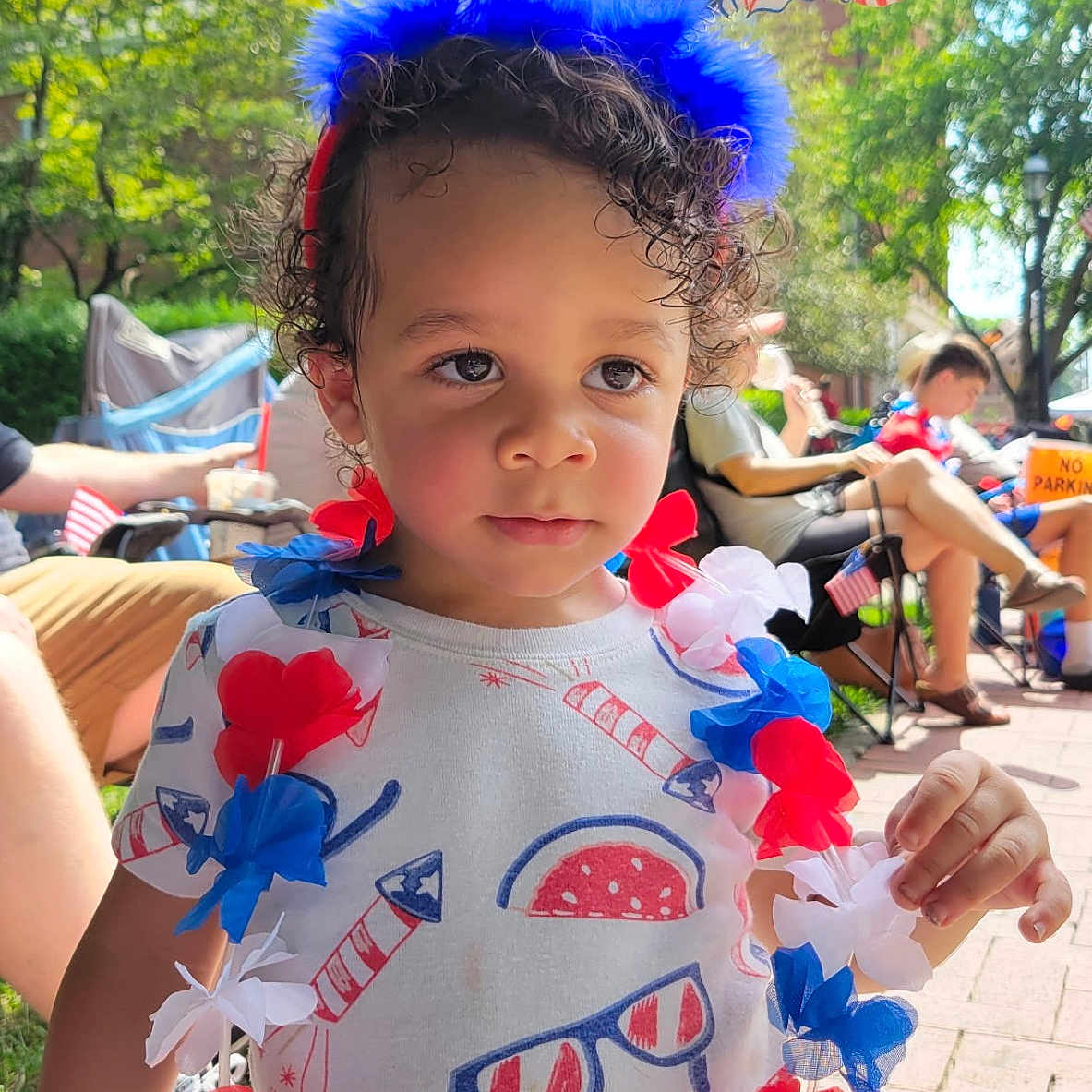 Demerious is registered to the contest to win money with this photo: toddler, child, outdoor, party, festive, headband, lei, red_white_blue, curly_hair, summer, celebration, people, sitting, grass, sidewalk, daylight, patriotic, balloons, clothing, portrait