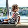 Demerious is registered to the contest to win money with this photo: toddler, child, curly_hair, smiling, window_seat, cityscape, sunlight, shorts, shirt, footwear, indoor, daylight, glass_window, urban_view, happy, casual_clothing, young_child, person, portrait, sitting