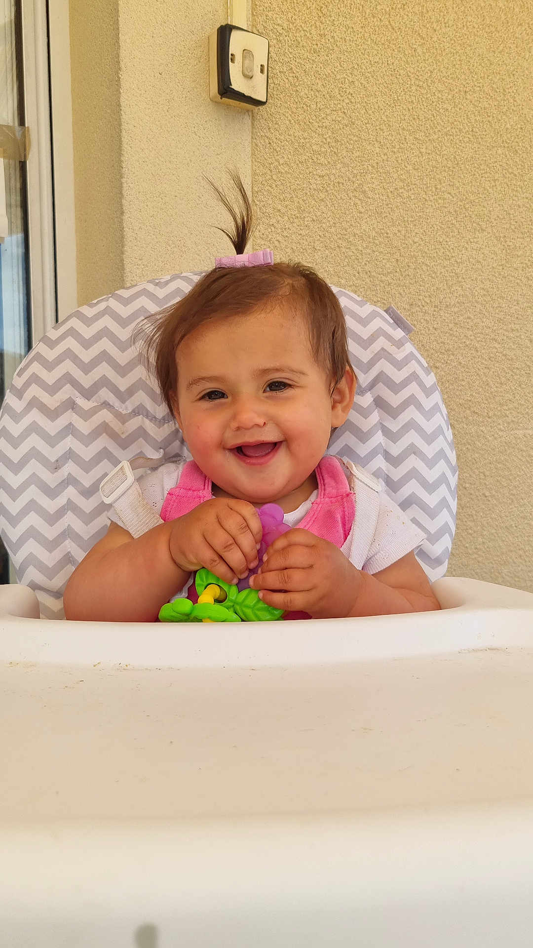 Ilyana participe au concours pour gagner de l'argent avec cette photo : baby, child, smiling, high_chair, toy, pink_clothing, ponytail, indoor, happy, face, person, seat, window, hand, white, skin, hair, playful, infant, cute