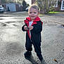 toddler, child, costume, black, red, standing, outdoor, daylight, smile, holding_object, rock, pavement, shadow, building, tree, grass, shoes, person, face, cute
