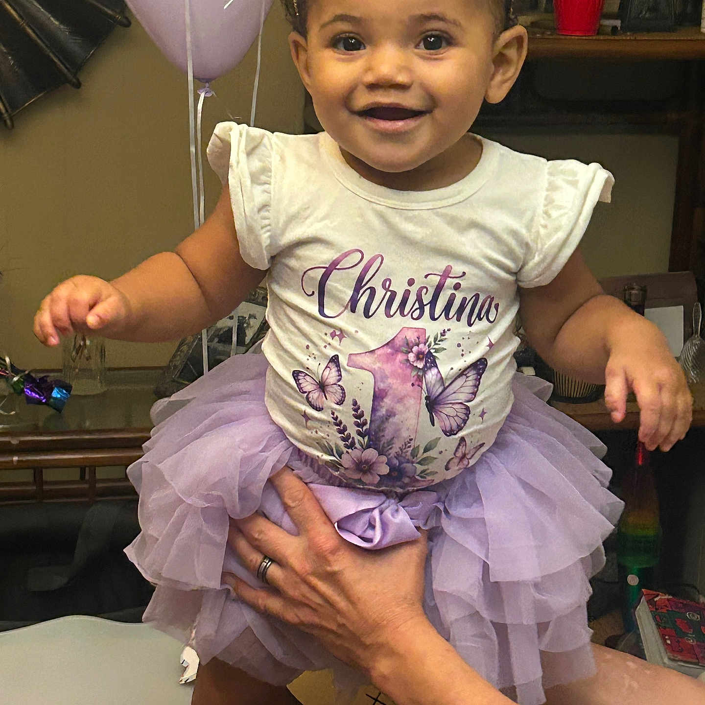 Christina is registered to the contest to win money with this photo: toddler, child, smiling, portrait, tiara, dress, tutu, lavender, butterflies, birthday, balloons, indoor, celebration, sitting, being_held, adult_hands, family, happy, furniture, home