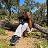 black_dog, blue_sky, casual_clothing, daytime, dog, fallen_tree, forest, grass, ground, long_hair, nature, outdoor, pet, plants, relaxing, shadows, sneakers, sunlight, tree, woman