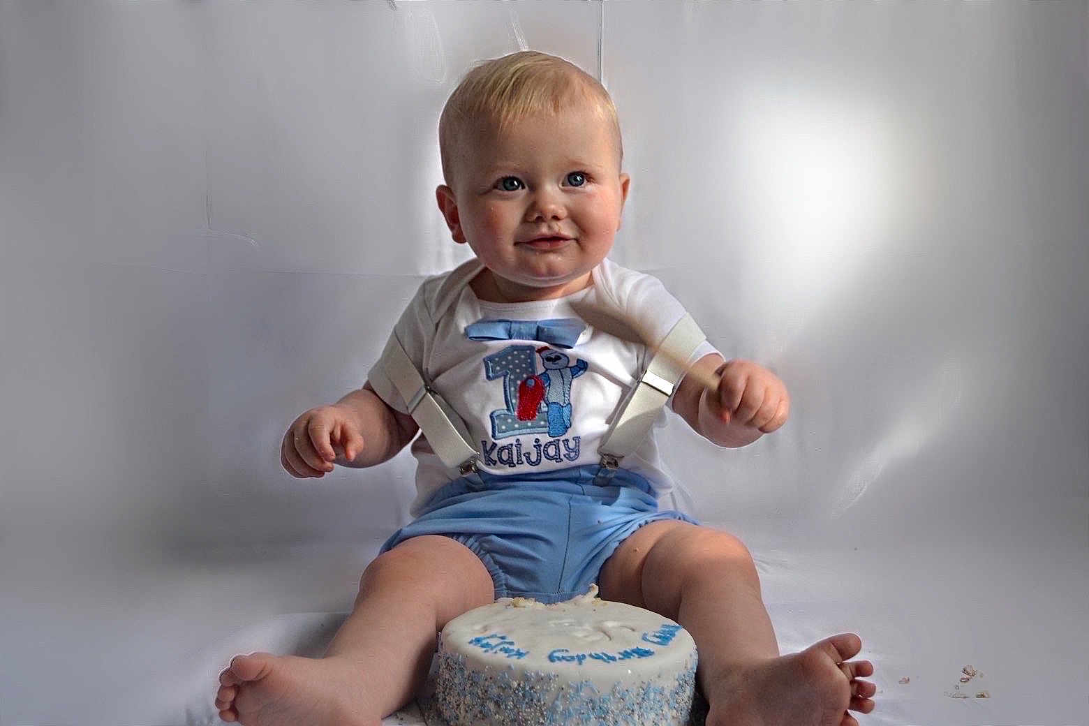 Kaijay joined the competition — help win amazing prizes! baby, child, birthday_cake, cake, blue_shorts, suspenders, smiling, sitting, first_birthday, celebration, cute, portrait, indoor, light_background, happy, face, feet, hands, clothing, toddler