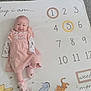 baby, infant, smiling, pink_dress, milestone_blanket, numbers, circled_number, lying_down, cartoon_characters, floor, soft_lighting, cute, child, footwear, happy, person, indoors, playful, blanket, age_marker