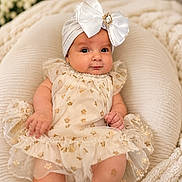 Yvonne joined the competition — help win amazing prizes! baby, infant, child, dress, headband, bow, cushion, soft_furniture, floral_pattern, cream_color, gold_details, portrait, indoors, cozy, cute, smiling, sitting, person, closeup, adorable