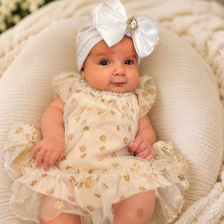 Yvonne joined the competition — help win amazing prizes! baby, infant, child, dress, headband, bow, cushion, soft_furniture, floral_pattern, cream_color, gold_details, portrait, indoors, cozy, cute, smiling, sitting, person, closeup, adorable