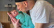 Harmony is registered to the contest to win money with this photo: baby, newborn, man, hospital_room, cap, white_tshirt, blanket, swaddled, holding, kissing, face, curious, love, affection, parent, indoor, medical_equipment, watch, chain, wood_paneling