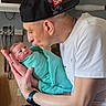 baby, newborn, man, hospital_room, cap, white_tshirt, blanket, swaddled, holding, kissing, face, curious, love, affection, parent, indoor, medical_equipment, watch, chain, wood_paneling