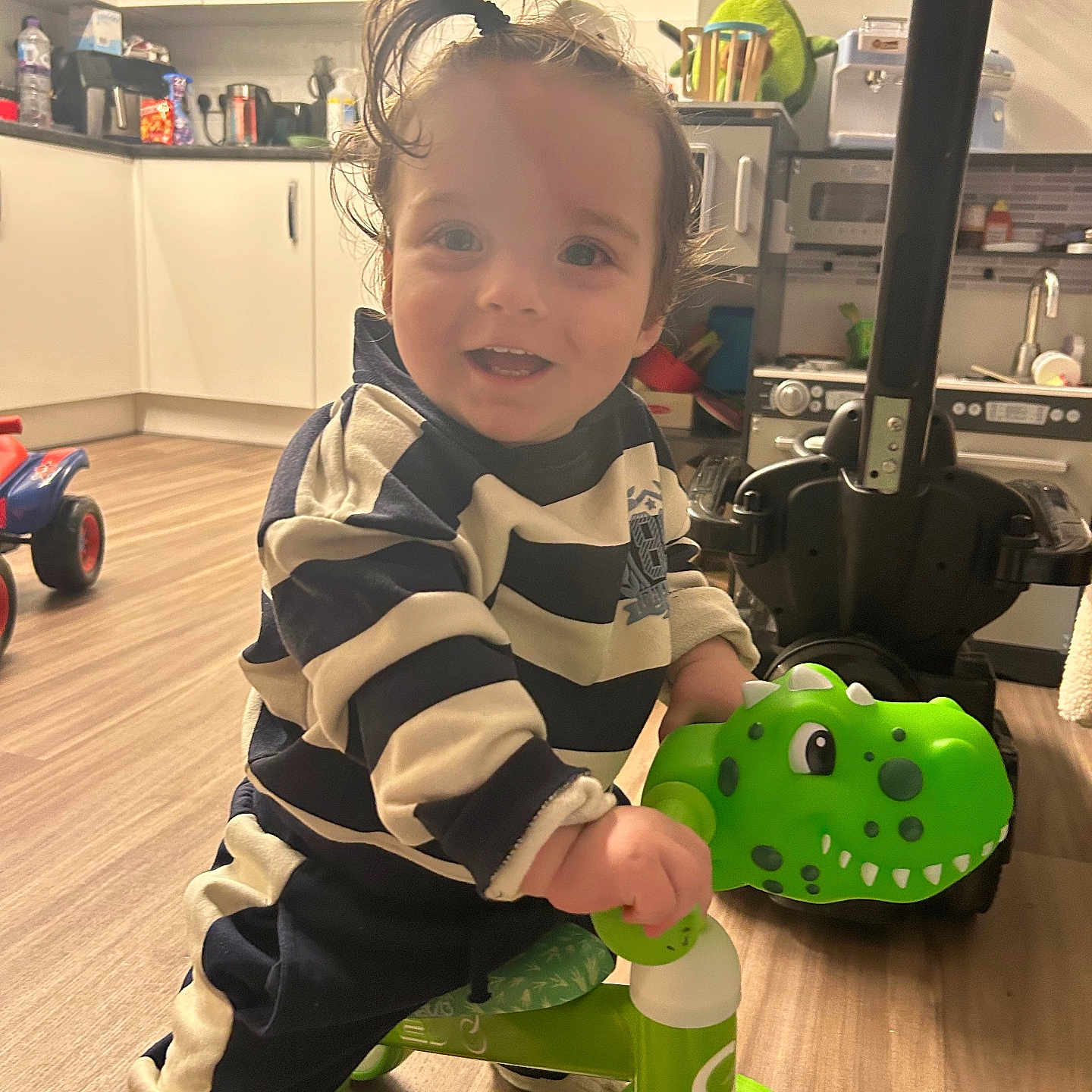 Gabriel is registered to the contest to win money with this photo: appliance, cabinet, child, clothing, dinosaur, floor, fun, hair, happy, indoor, kitchen, person, plastic_toy, playful, ride, smiling, toddler, toy, tricycle, young_child