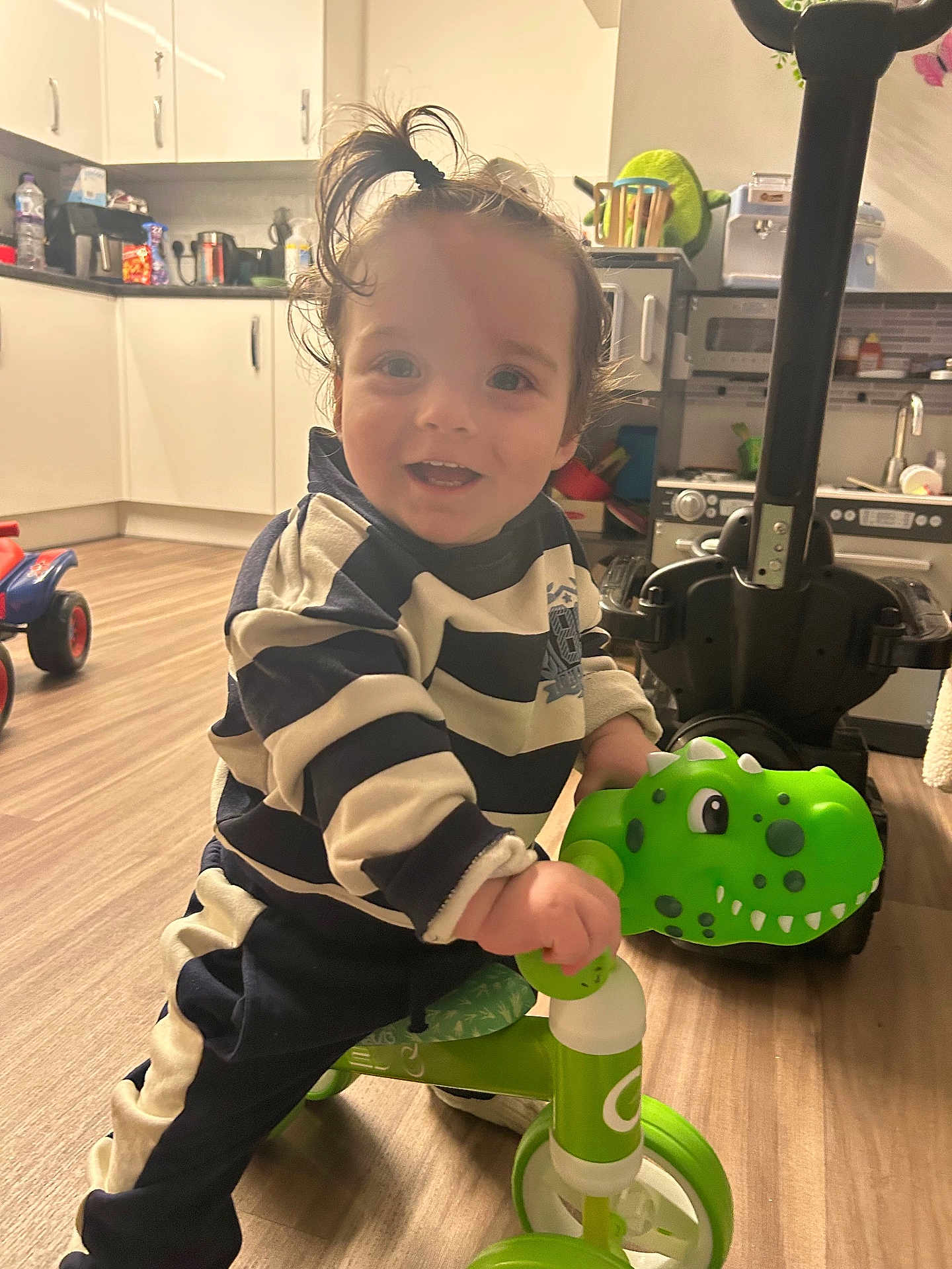 Gabriel is registered to the contest to win money with this photo: toddler, child, indoor, toy, tricycle, dinosaur, smiling, playful, hair, kitchen, floor, cabinet, appliance, person, young_child, happy, clothing, plastic_toy, fun, ride