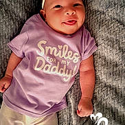 Oni is registered to the contest to win money with this photo: baby, infant, smiling, tongue_out, headband, purple_shirt, text_on_clothing, blanket, soft_texture, lying_down, cute, happy, child, person, casual_clothing, white_socks, playful, indoors, portrait, closeup
