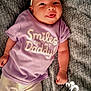 baby, infant, smiling, tongue_out, headband, purple_shirt, text_on_clothing, blanket, soft_texture, lying_down, cute, happy, child, person, casual_clothing, white_socks, playful, indoors, portrait, closeup