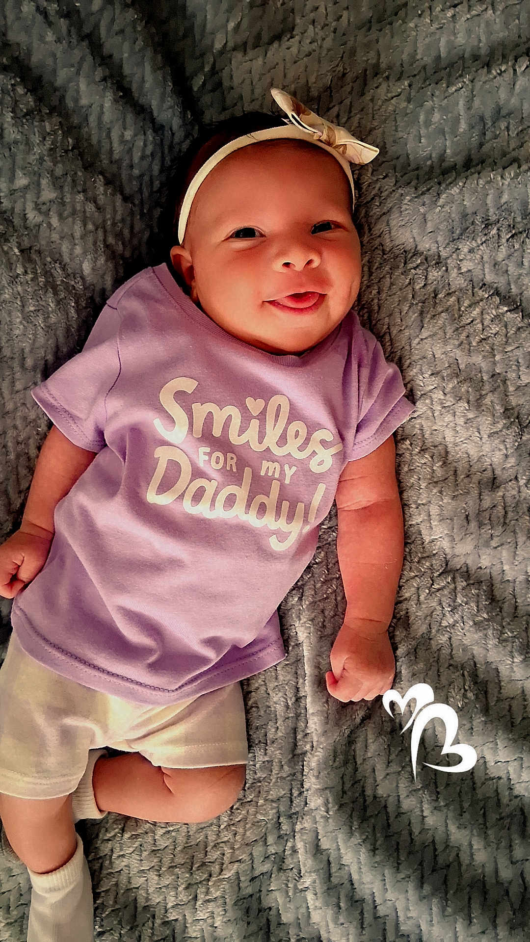 Oni is registered to the contest to win money with this photo: baby, infant, smiling, tongue_out, headband, purple_shirt, text_on_clothing, blanket, soft_texture, lying_down, cute, happy, child, person, casual_clothing, white_socks, playful, indoors, portrait, closeup