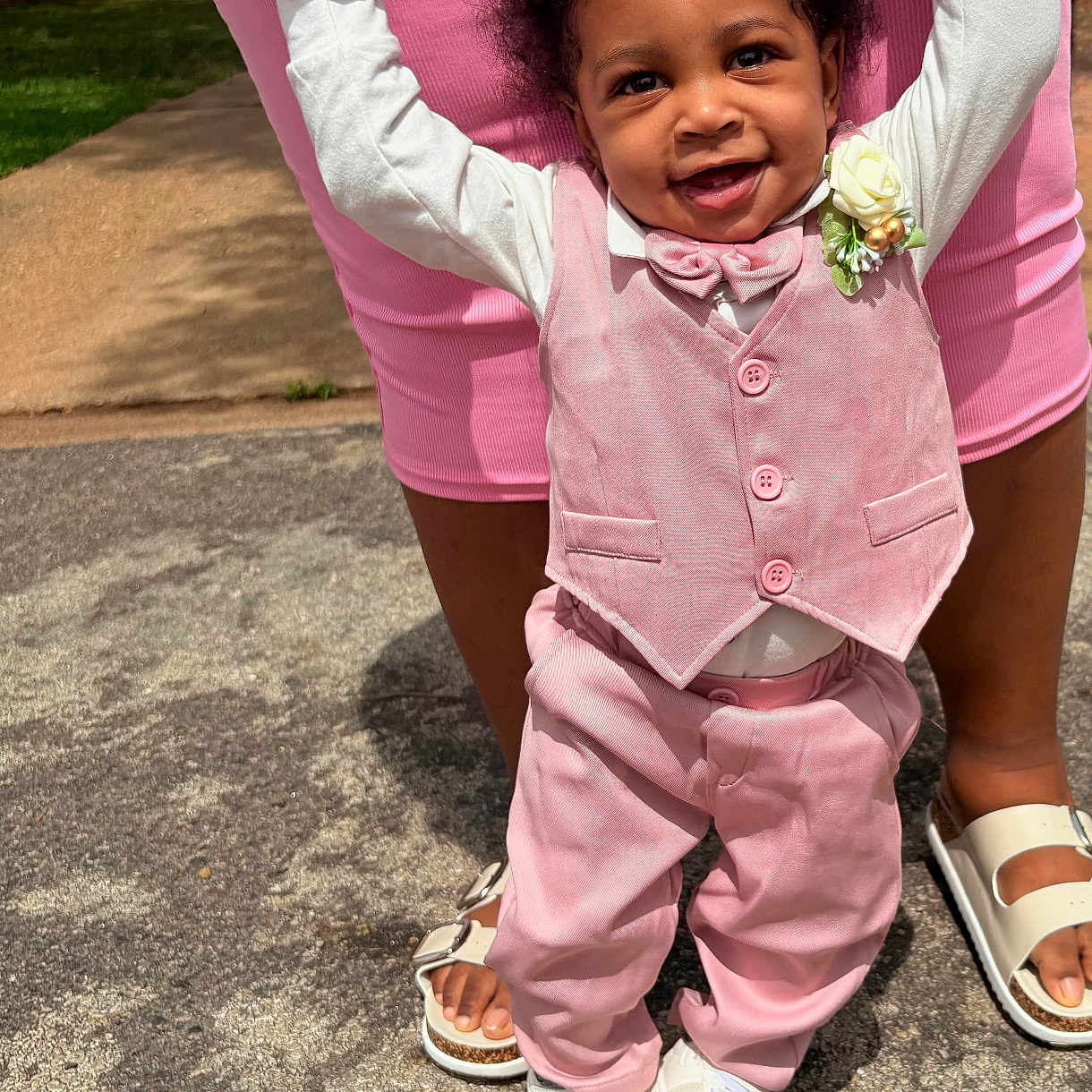 Trevon is registered to the contest to win money with this photo: baby, child, smiling, pink_clothing, vest, bow_tie, flower_corsage, white_shoes, adult_hands, supporting, outdoors, pavement, sunlight, happy, person, standing, cute, fashion, footwear, grass