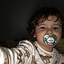 toddler, child, face, curly_hair, pajamas, dinosaur_pattern, pacifier, selfie, indoor, low_light, person, young_child, cute, baby, sleepwear, hand, portrait, expression, closeup, childhood