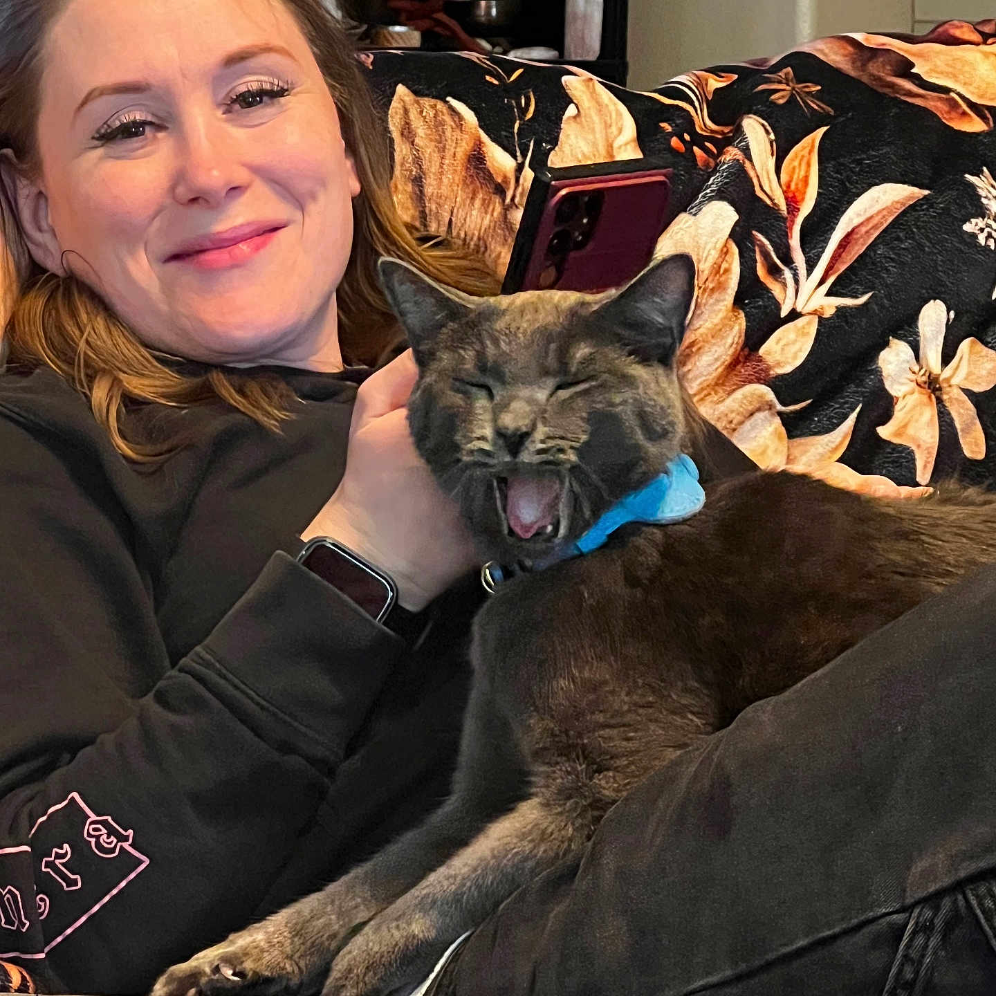 Louie is registered to the contest to win money with this photo: black_sweatshirt, blanket, cat, collar, comfort, couch, face, fur, gray_cat, hand, happy, indoor, patterned_blanket, pet, relaxed, resting, smartwatch, smiling, woman, yawning