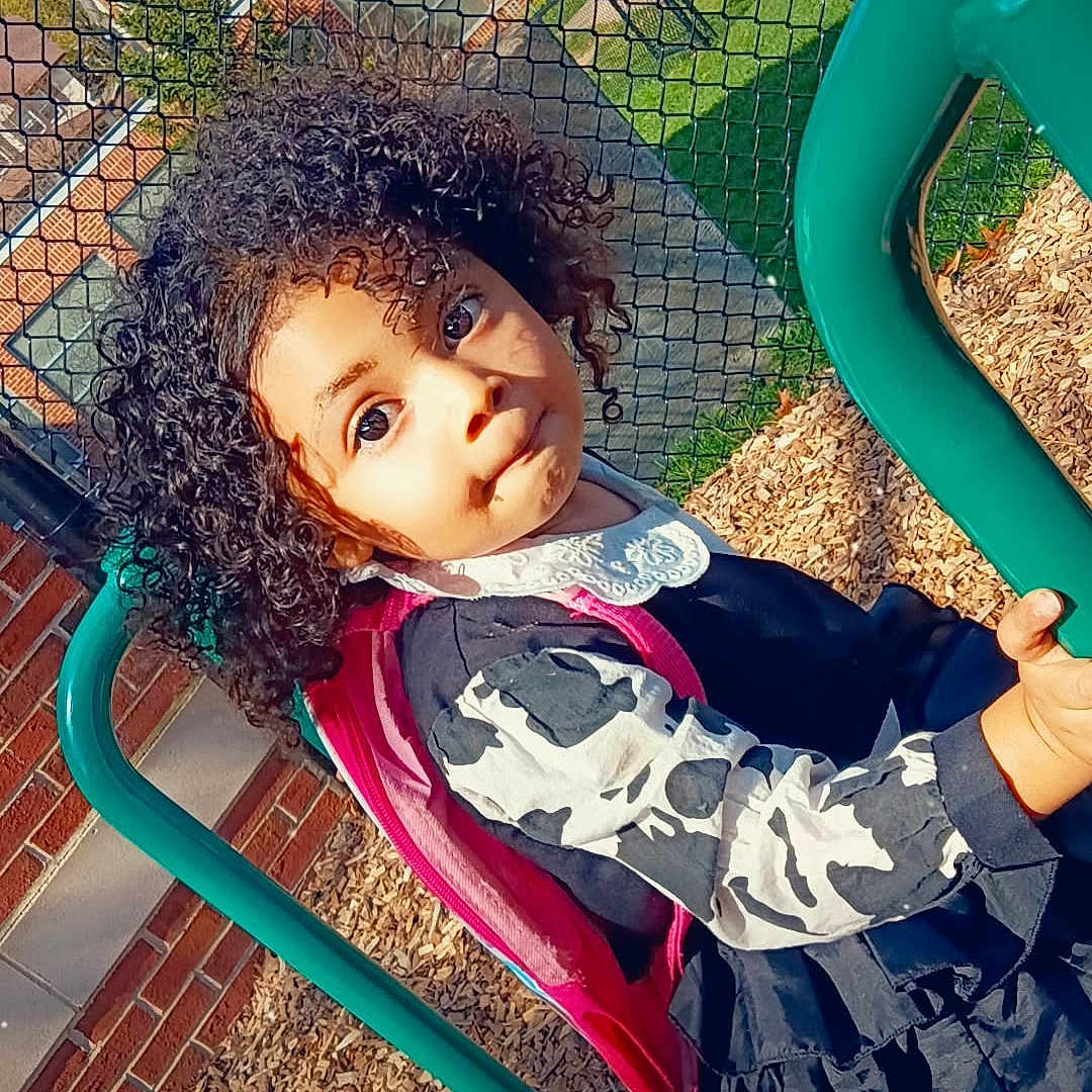 Ashanti Singleton is registered to the contest to win money with this photo: child, girl, curly_hair, backpack, playground, green_bars, outdoor, fence, brick_wall, wood_chips, daylight, casual_clothing, expression, person, young, cute, park, recreation, smile, head