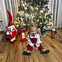 child, toddler, christmas_tree, holiday_decorations, gnome, lantern, wooden_floor, smiling, sitting, indoors, festive, lights, ornaments, red_clothing, white_clothing, curtains, home, celebration, happy, toy_car