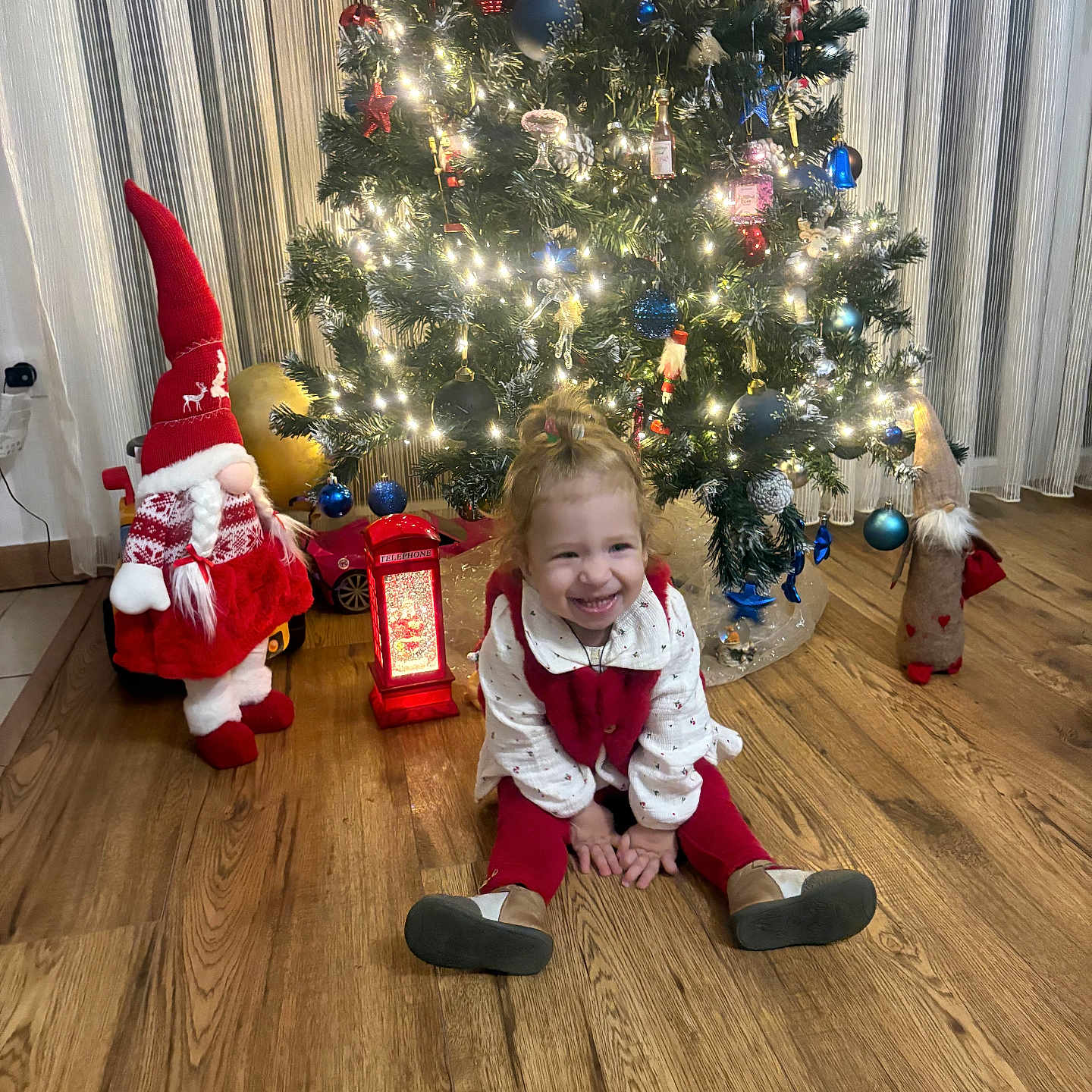 Tamari participe au concours pour gagner de l'argent avec cette photo : celebration, child, christmas_tree, curtains, festive, gnome, happy, holiday_decorations, home, indoors, lantern, lights, ornaments, red_clothing, sitting, smiling, toddler, toy_car, white_clothing, wooden_floor