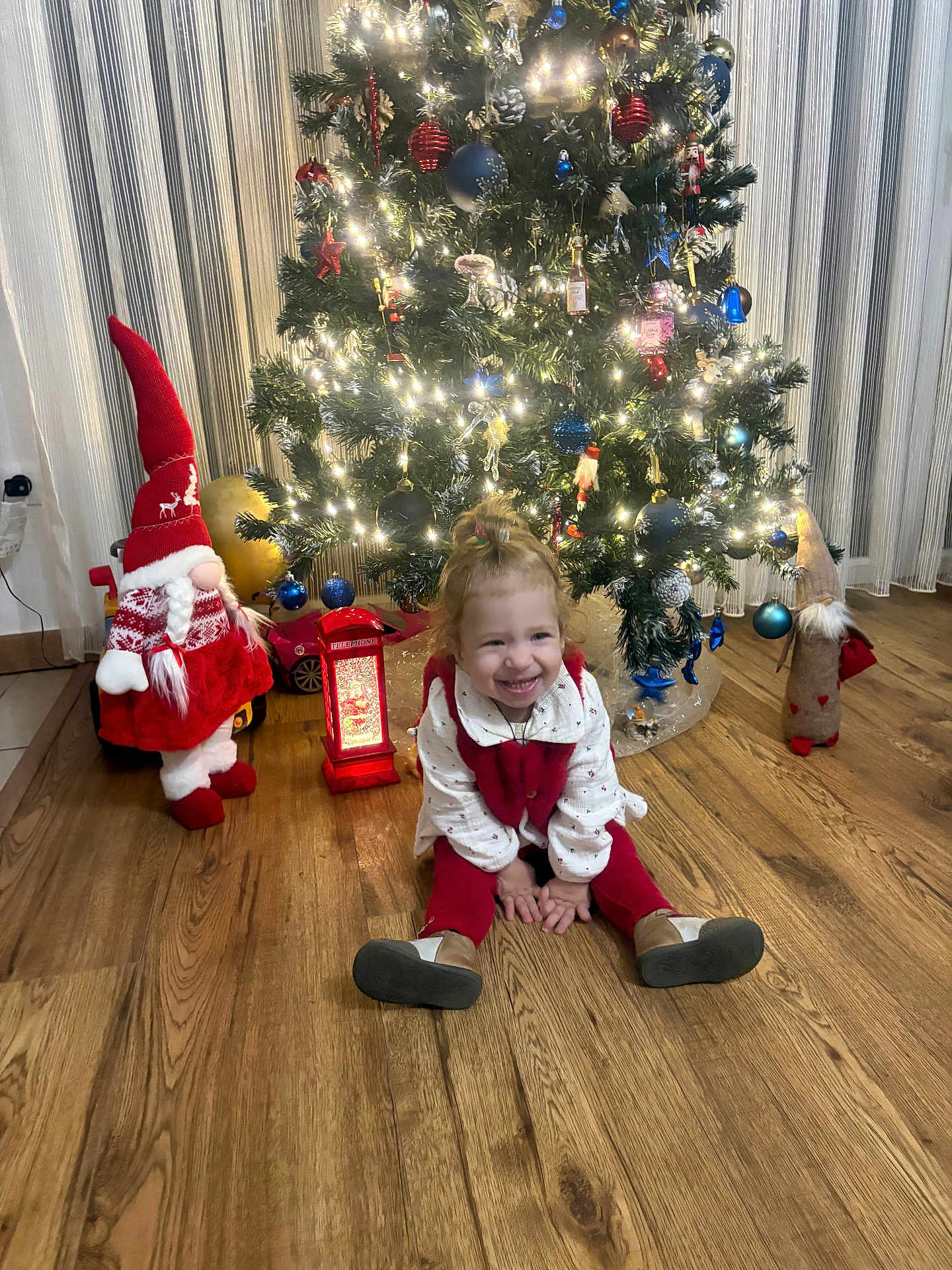 Tamari participe au concours pour gagner de l'argent avec cette photo : child, toddler, christmas_tree, holiday_decorations, gnome, lantern, wooden_floor, smiling, sitting, indoors, festive, lights, ornaments, red_clothing, white_clothing, curtains, home, celebration, happy, toy_car