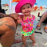toddler, child, beach, sand, sunhat, pink_hat, swimsuit, colorful_clothing, smiling, happy, tattoo, adult_leg, outdoor, summer, vacation, umbrella, barefoot, person, sunny, fun