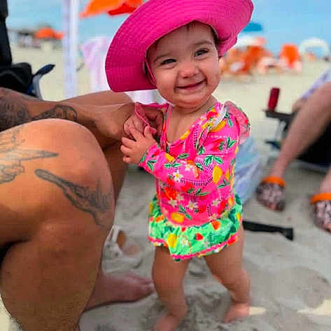 Stephanie Gonzaga is registered to the contest to win money with this photo: toddler, child, beach, sand, sunhat, pink_hat, swimsuit, colorful_clothing, smiling, happy, tattoo, adult_leg, outdoor, summer, vacation, umbrella, barefoot, person, sunny, fun